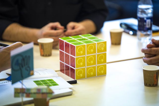 A cube with green, red and yellow faces on a meeting table, between participants’ hands